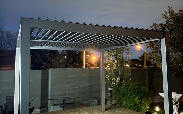 A modern louvered pergola installed on a wooden deck at night.