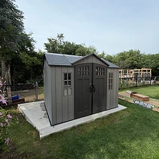 A newly assembled grey storage shed stands on a concrete foundation in a residential backyard.