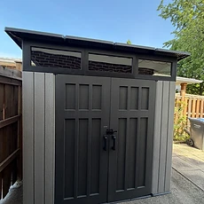 A dark gray resin shed with windows installed on a driveway pad.