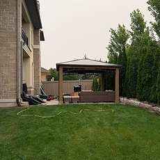 A gazebo with curtains is installed on a residential deck near a brick house.