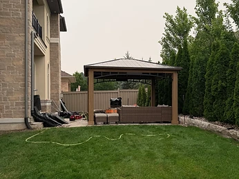 A gazebo with curtains is installed on a residential deck near a brick house.