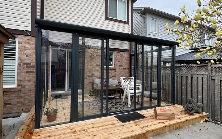 A glass and black frame solarium installation sits on a raised wooden deck attached to a brick home.