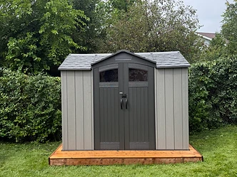 A grey resin shed is installed on a wooden base in a backyard with green grass.