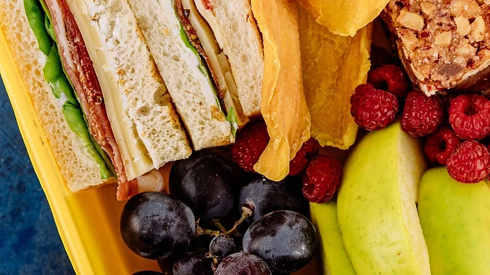A packed lunchbox with sandwiches, grapes, raspberries, apple slices and a cereal bar