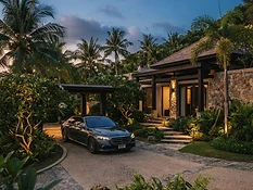 A discreet chauffeured luxury sedan waiting on a palm-lined villa driveway at dusk