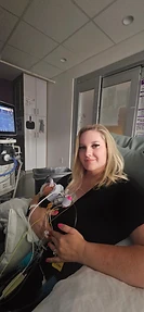 A mother holding her premature daughter skin-to-skin in a NICU chair, surrounded by monitors and medical tubing.