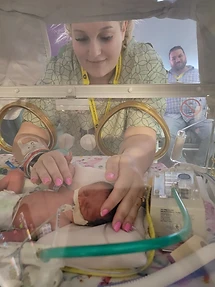A mother leaning into a NICU isolette, gently placing her hands on her premature baby's chest and head, while a father watches nearby.