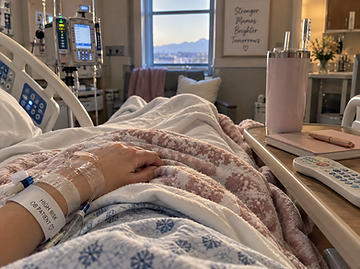 A first-person view from a hospital bed of a patient's hand with an IV and a High Risk OB Patient wristband, resting on a pink throw blanket beside a tray table with a tumbler and notebook, looking out a window at a cityscape.