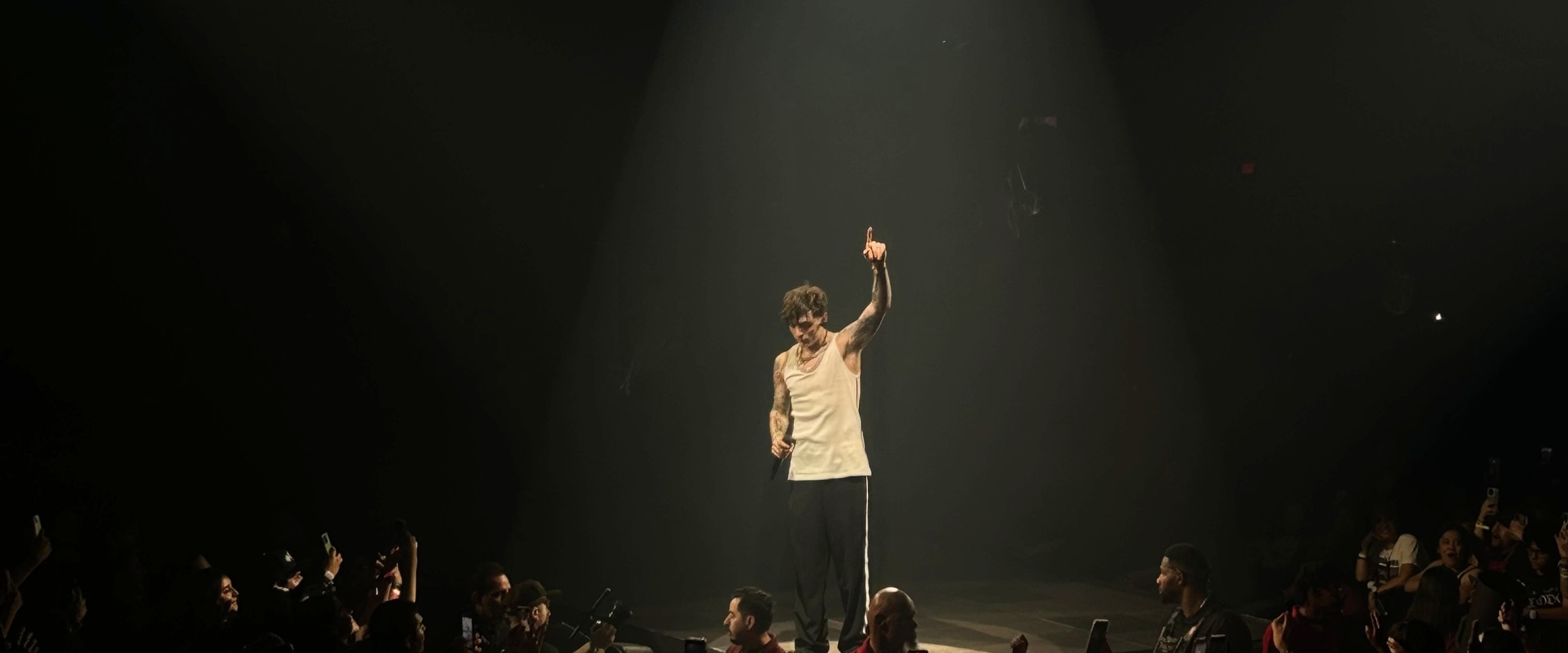 Peso Pluma on stage in a spotlight with one arm raised and fans around the stage
