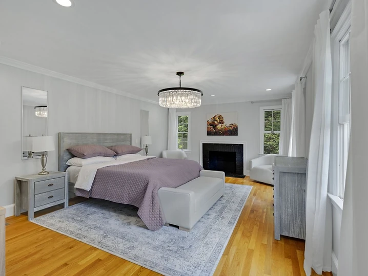 The primary bedroom with a lavender coverlet, a crystal chandelier, and a black fireplace flanked by white chairs