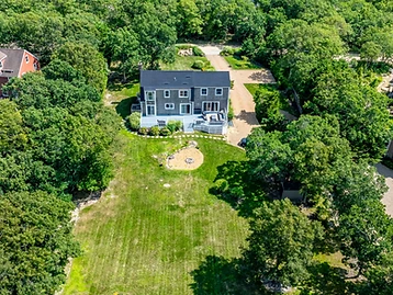 Top-down aerial of the shingled house, deck, fire pit lawn, and gravel driveway ringed by mature forest