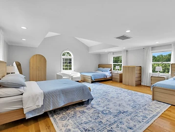 Vaulted bedroom with three light-blue beds, arched window, white armchairs, and light oak wardrobe
