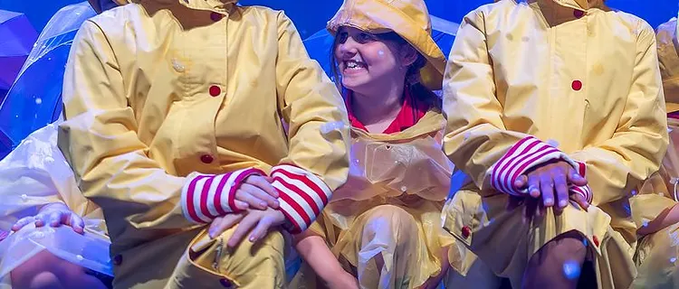 Performers in yellow rain slickers dancing in falling 'rain' under blue stage lights