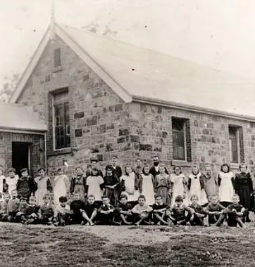 Historical group photograph of students and teacher in front of the stone school building