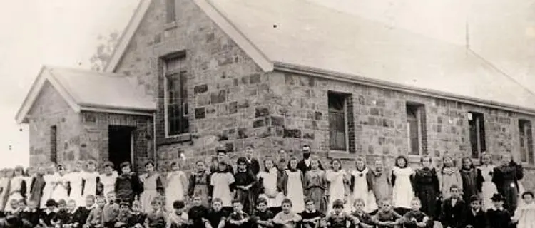 Historical photograph of students and teacher in front of the stone school building