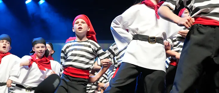 Performers in pirate costumes with red sashes singing on stage under blue spotlights