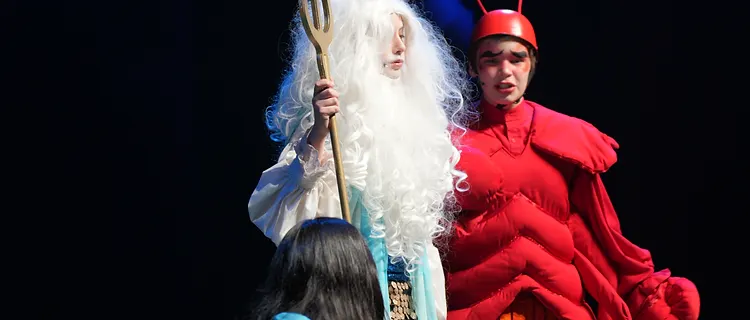 Performers in mermaid, trident and crab costumes under stage spotlights