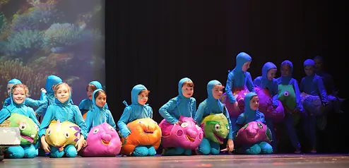 Students in blue costumes carrying colourful plush fish across a lit stage