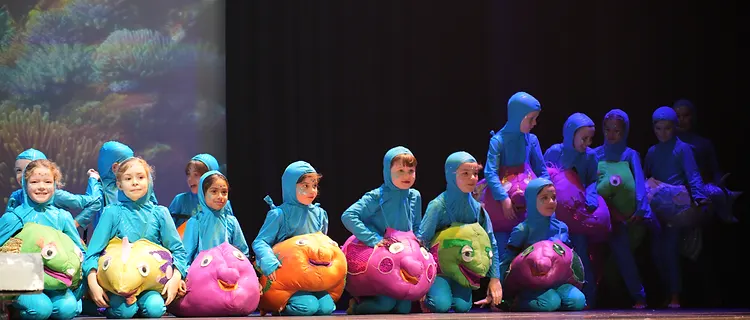 Students in blue costumes carrying colourful plush fish across a lit stage