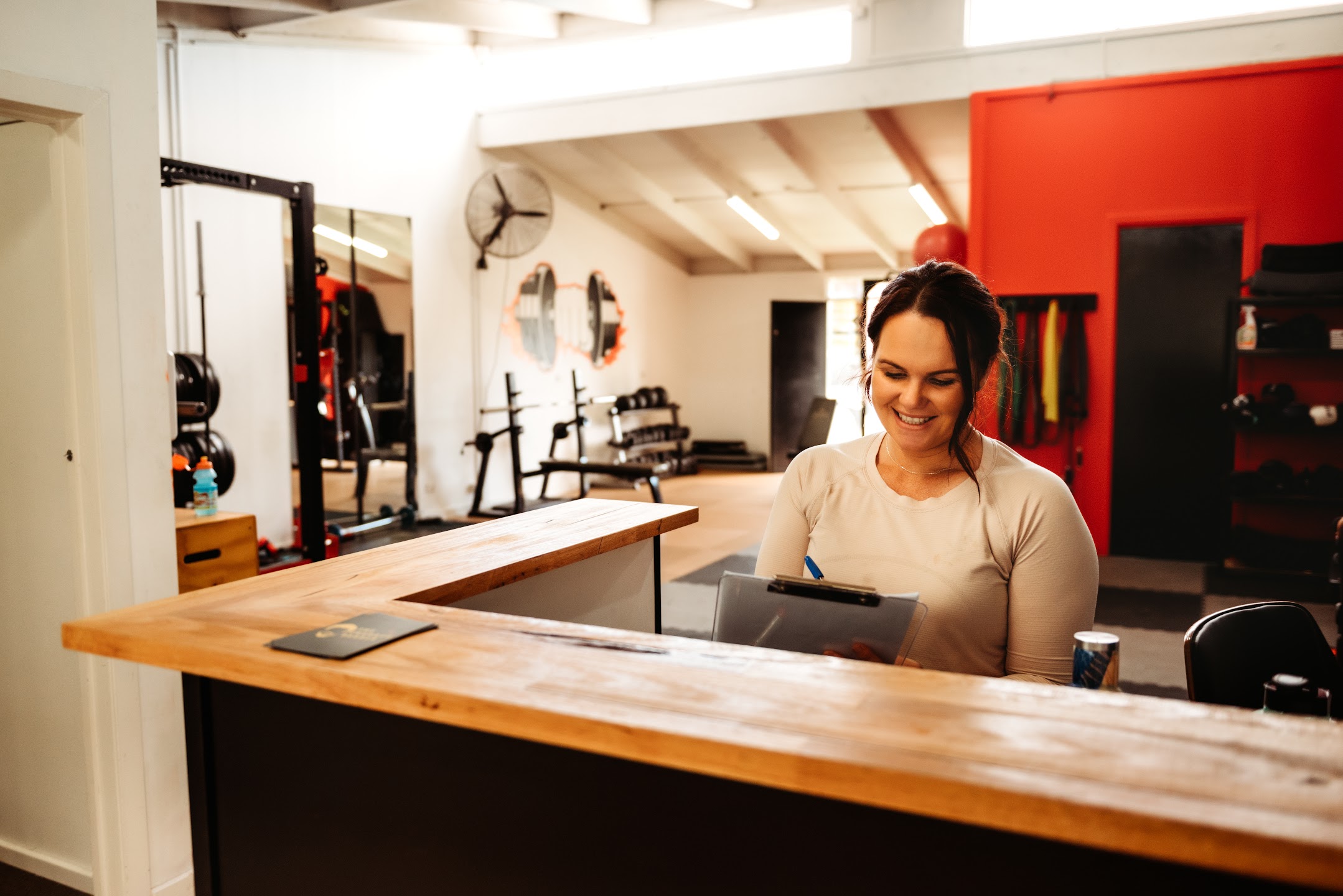 A smiling Familia Fitness staff member standing behind a rustic wooden reception desk, writing on a clipboard inside the gym facility