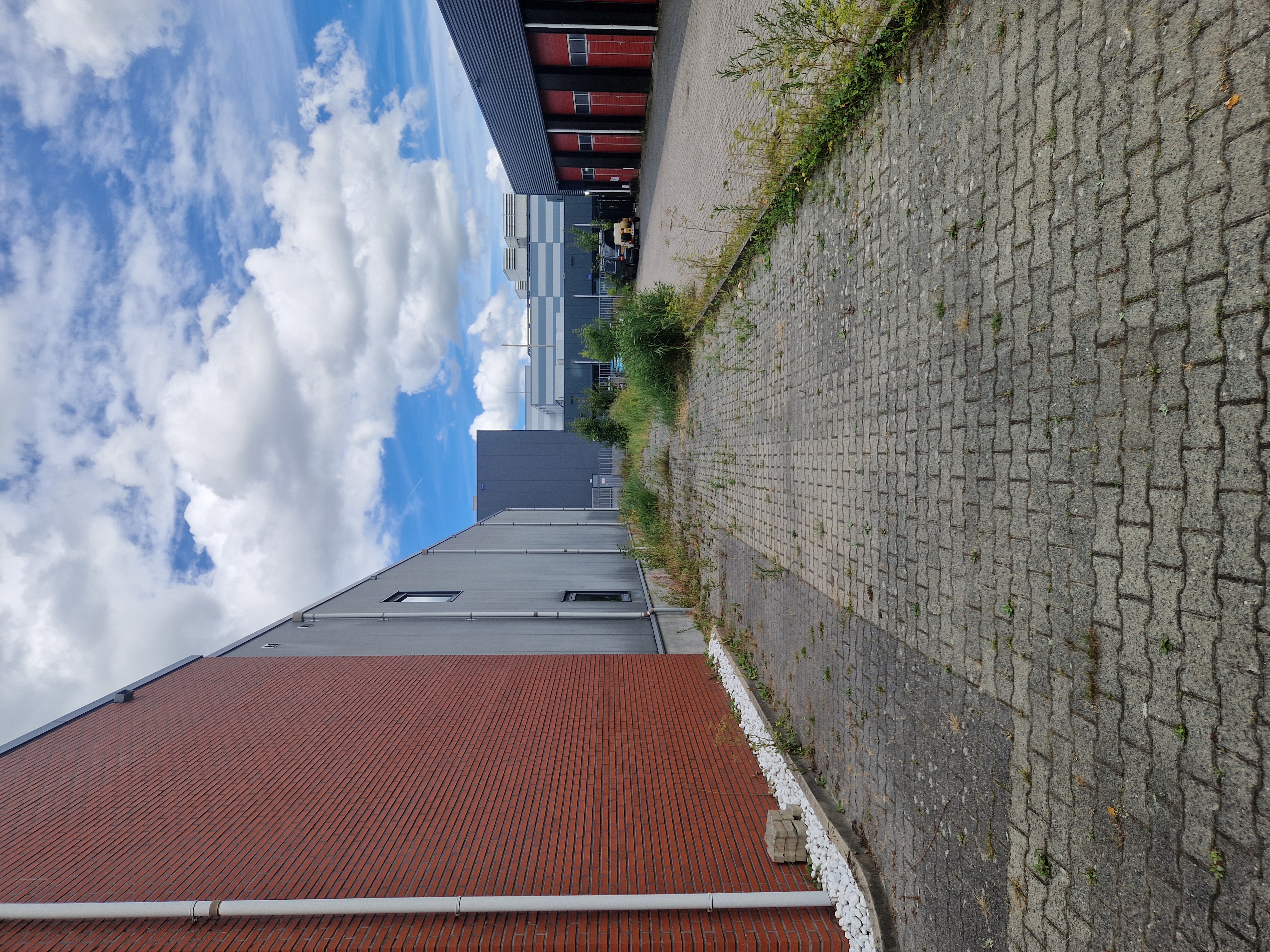 Zijaanzicht van een grijs industriegebouw met rode bakstenen plint, met onkruid dat tussen de bestrating groeit