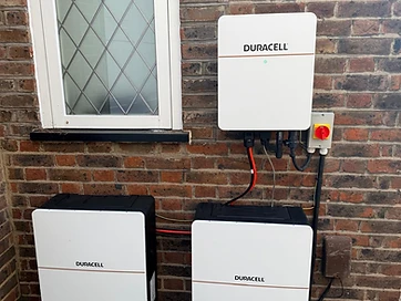 White Duracell inverter and two battery modules mounted on a dark brick wall beside a rotary isolator switch