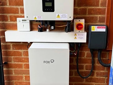 Fox ESS inverter with LCD screen and battery storage unit mounted on a red brick exterior wall with an isolator switch