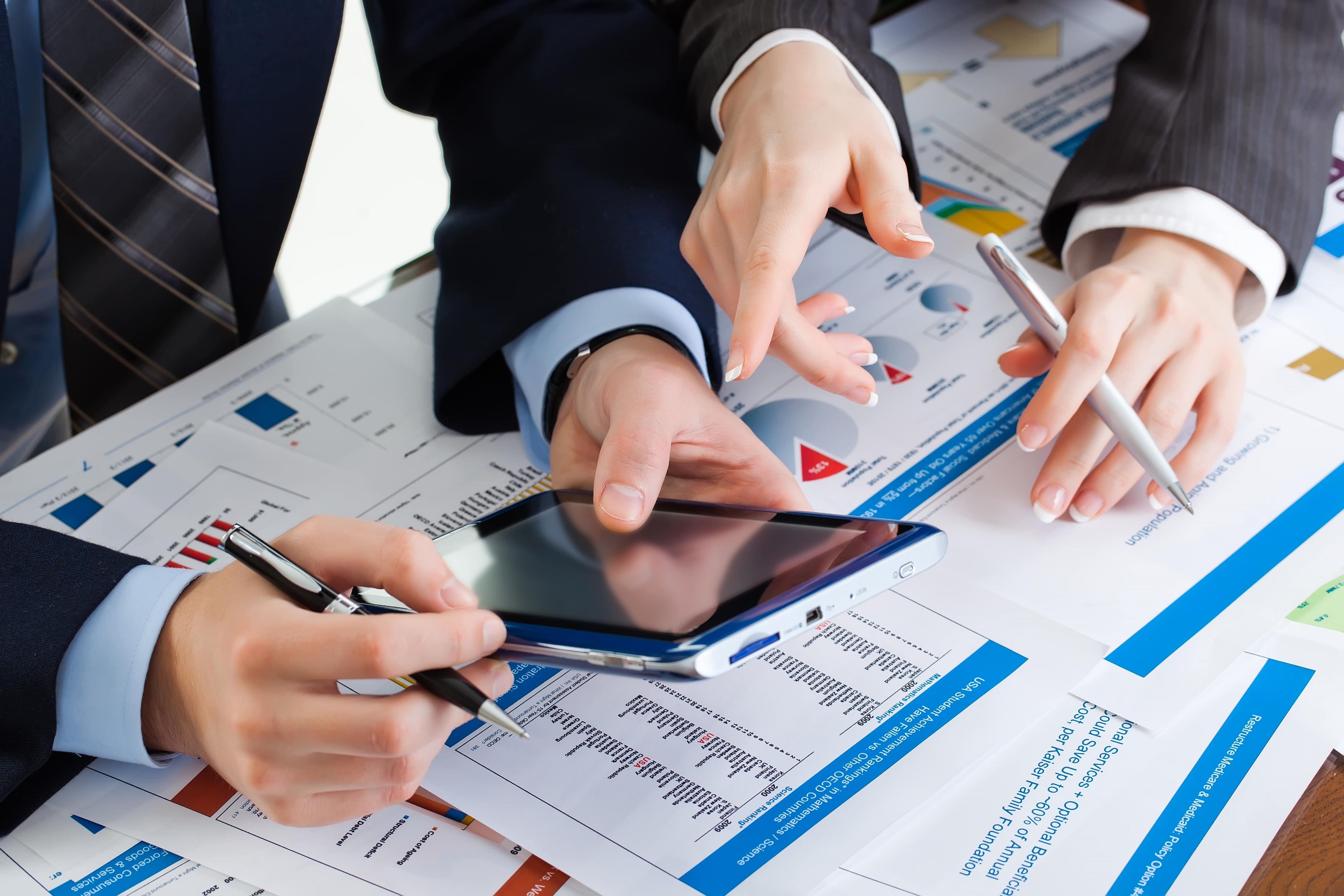 Two executives in business attire collaborating over a tablet and financial data reports, analysing fiscal charts and institutional policy documents in an office setting