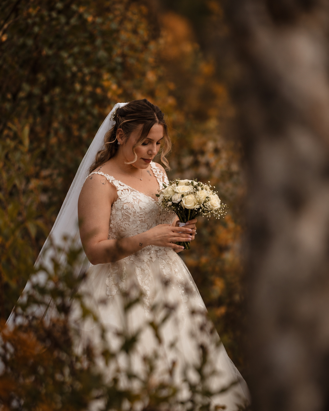 A bride looking down at her bouquet of white roses, a long sheer veil flowing behind her in an autumn forest