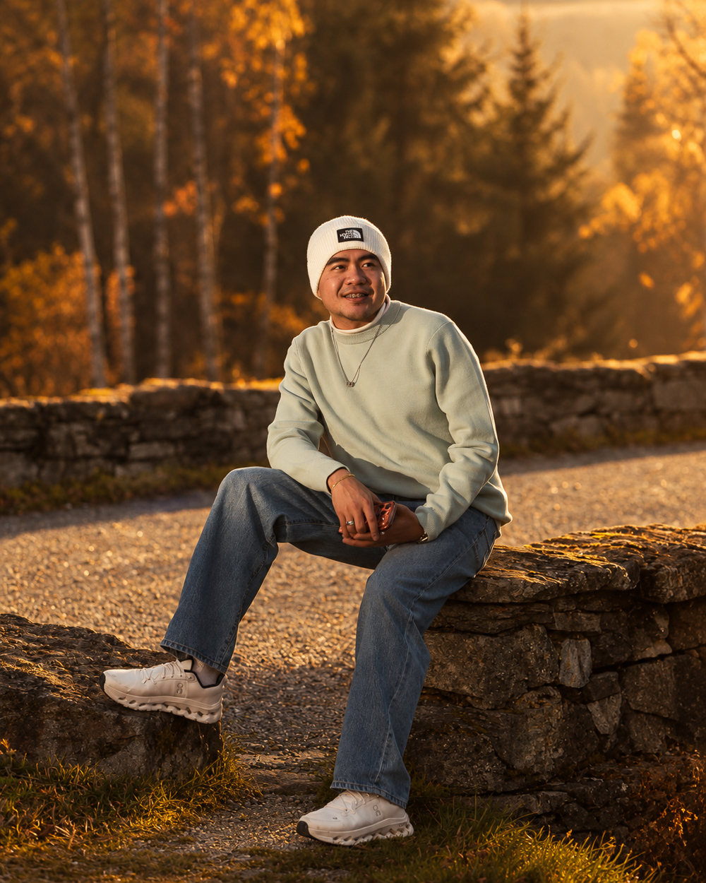 A young man in a white beanie and mint sweater sitting on a stone wall in a golden autumn forest