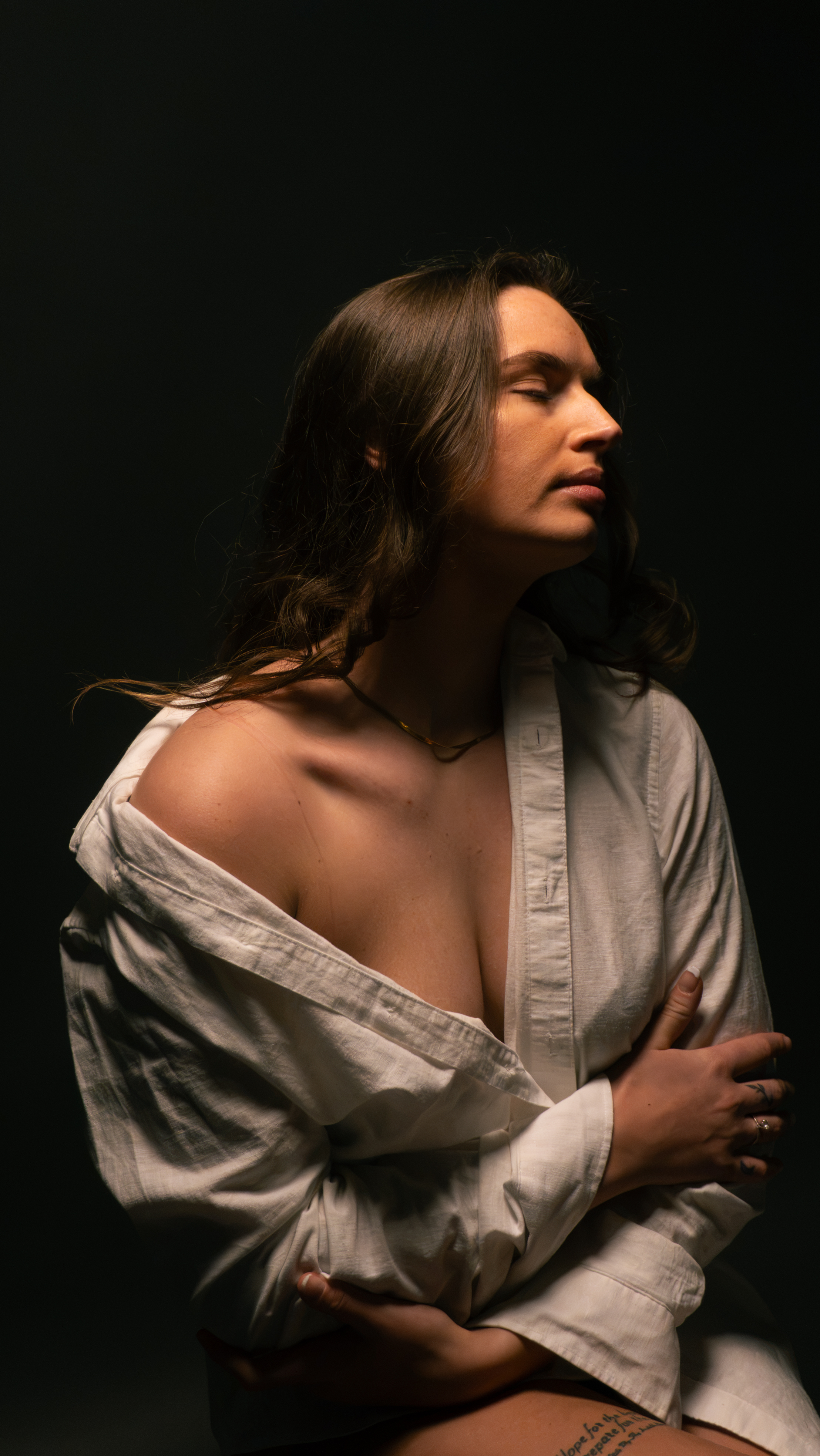 A woman in an oversized white linen shirt with eyes closed and arms folded, lit warmly from the upper right against a black studio backdrop