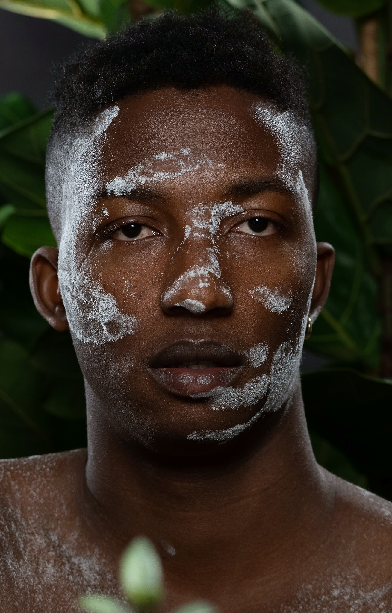 Portrait of a person with textured white powder on the face, set against dark green foliage