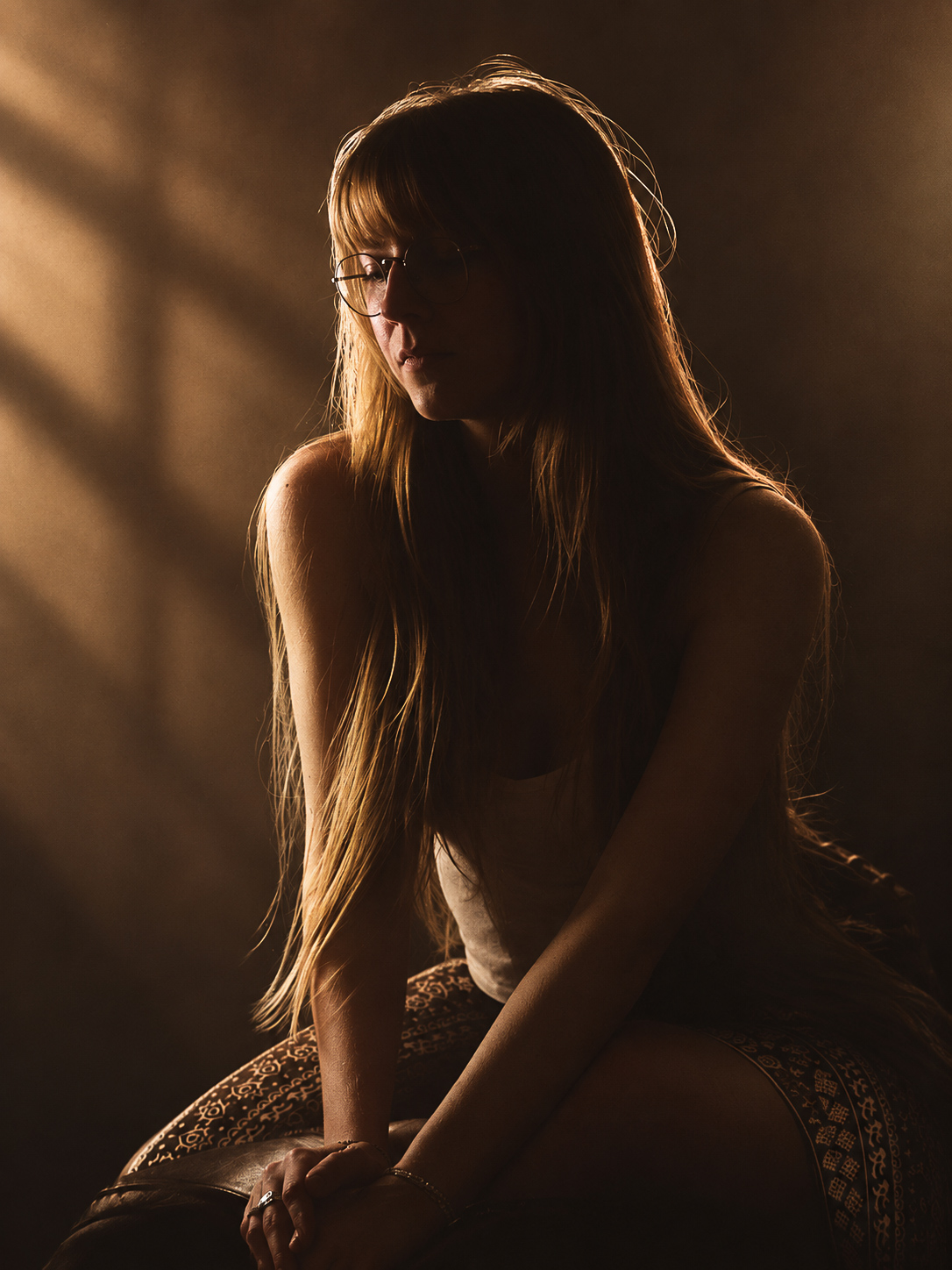 A young woman with long light-brown hair and round glasses seated in contemplation, hands clasped in her lap, bathed in warm slatted light and deep shadow