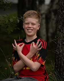A smiling boy in a red soccer jersey seated on a rock outdoors