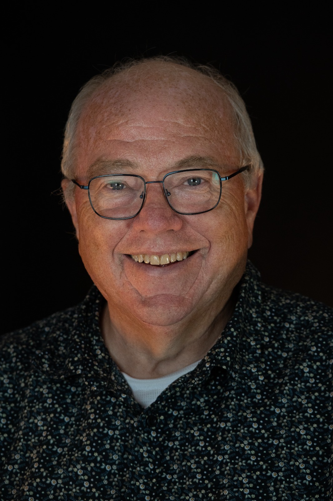 A warm portrait of an older man with glasses and a smile against a black backdrop