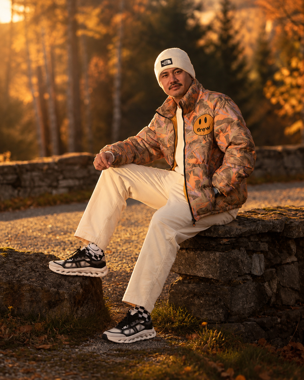 A young man in a cream beanie and patterned puffer jacket sitting on a stone wall in an autumn forest at sunset