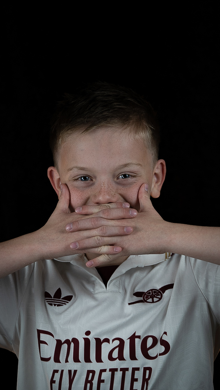A young boy in a white Arsenal soccer jersey with hands over his mouth against a black backdrop