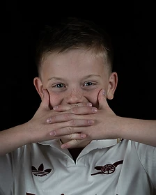 A young boy in a white Arsenal soccer jersey with hands over his mouth against a black backdrop