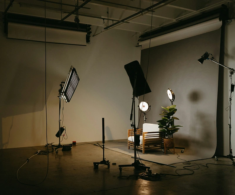 A photography studio set for a portrait shoot: a wooden armchair with white cushions on a grey paper backdrop, lit by a large softbox and studio lights