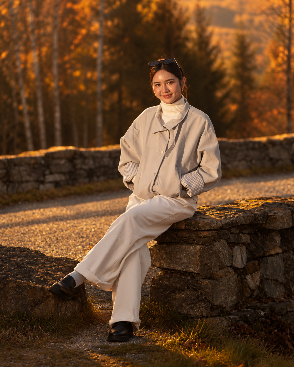 A young woman in a cream turtleneck and grey jacket sitting on a stone wall in warm golden-hour light