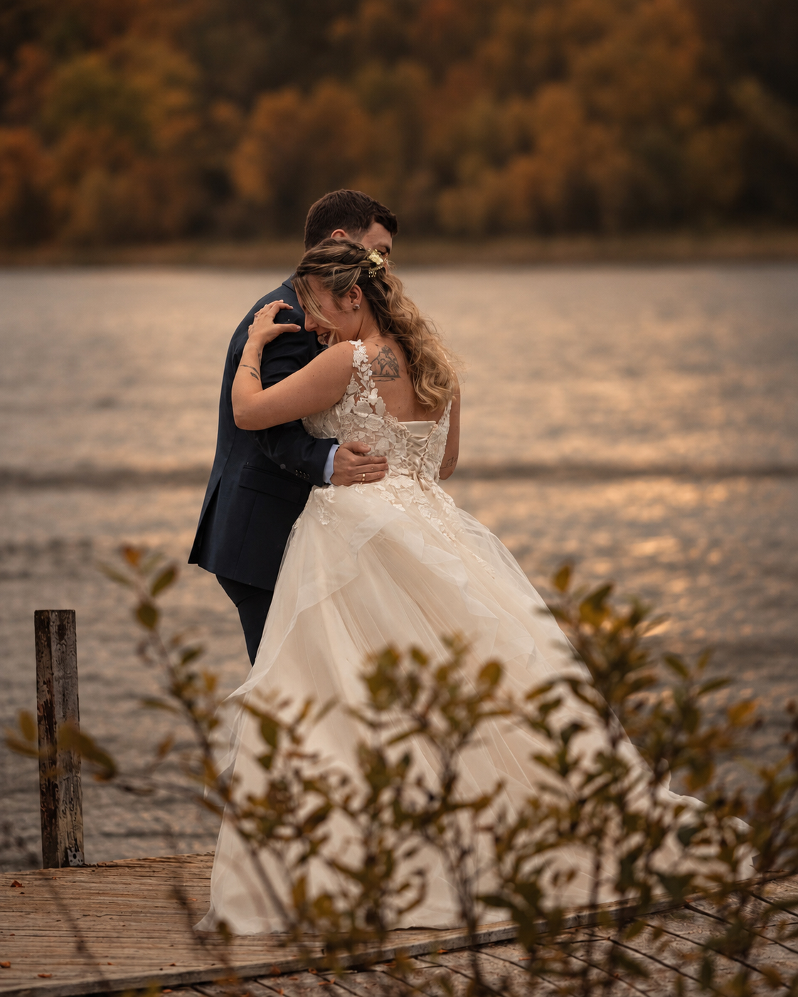 A bride and groom embracing on a wooden pier over calm water, autumn foliage reflected in the lake