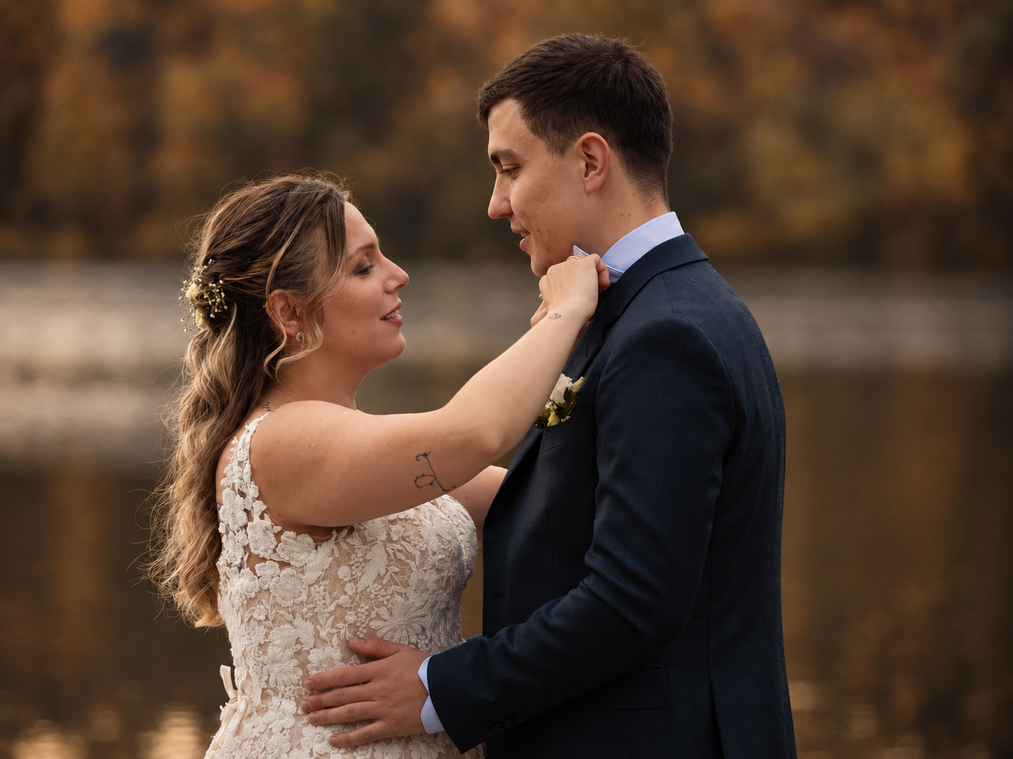 A bride adjusting her groom's bowtie by a lake in golden autumn light, both smiling gently