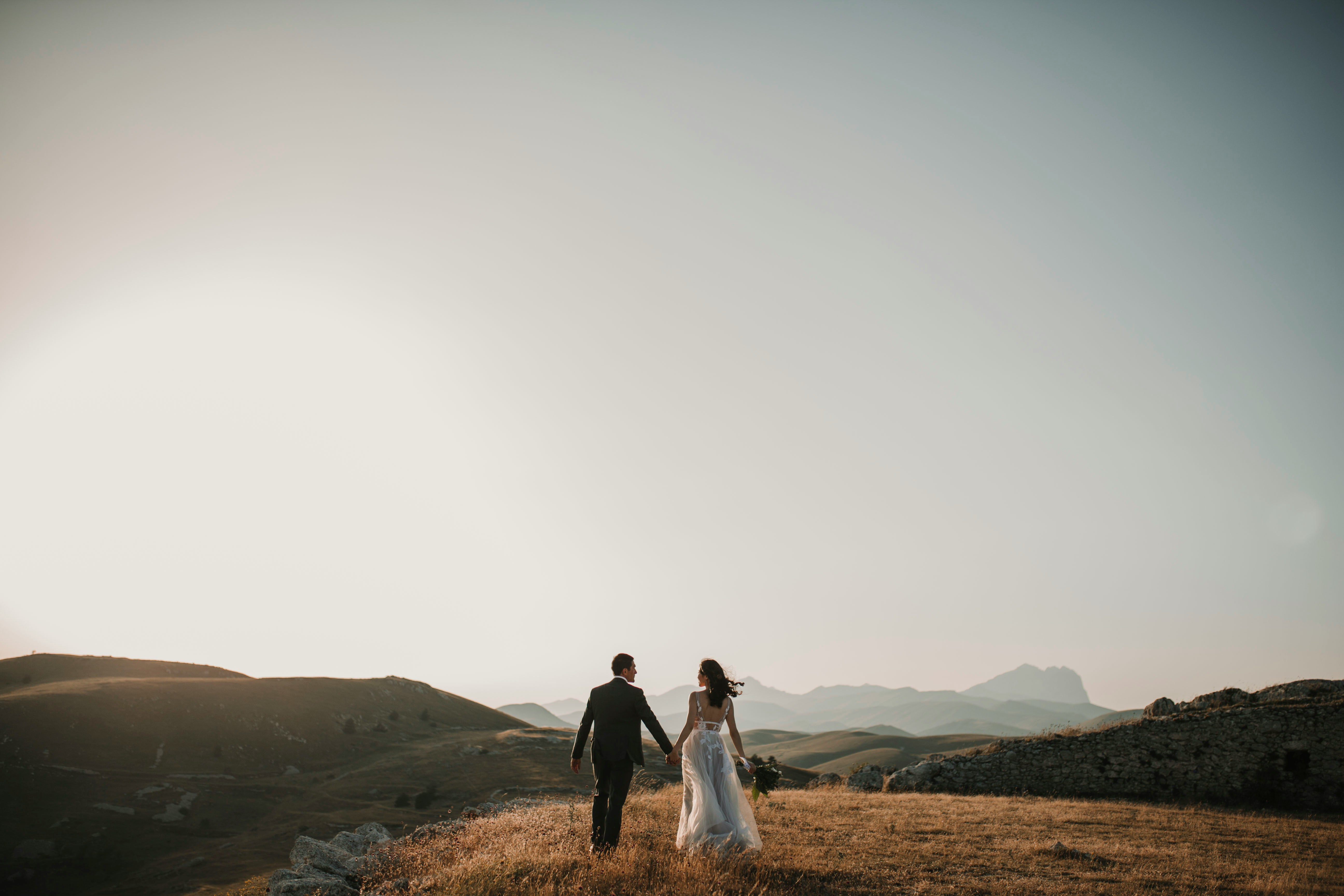 A bride and groom walking hand-in-hand across a golden meadow toward distant mountains