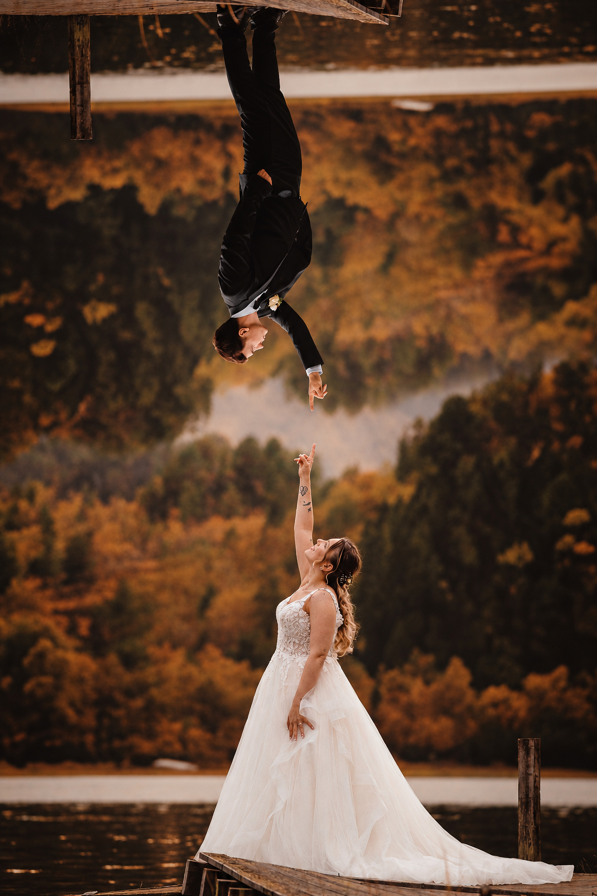 A surreal mirrored wedding portrait on a lake pier — the bride reaching up and the groom inverted above, fingertips nearly touching