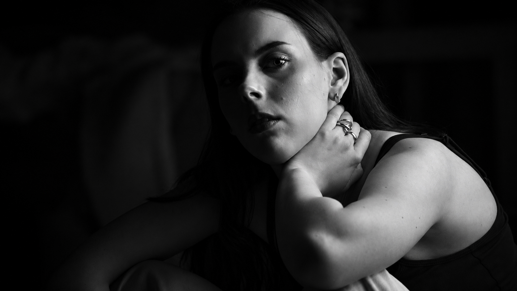 A young woman seated in a studio, hand resting against her neck, in high-contrast black and white