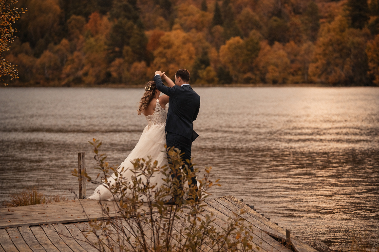 A newlywed couple dancing on a wooden pier over a calm lake, seen from behind in golden-hour light
