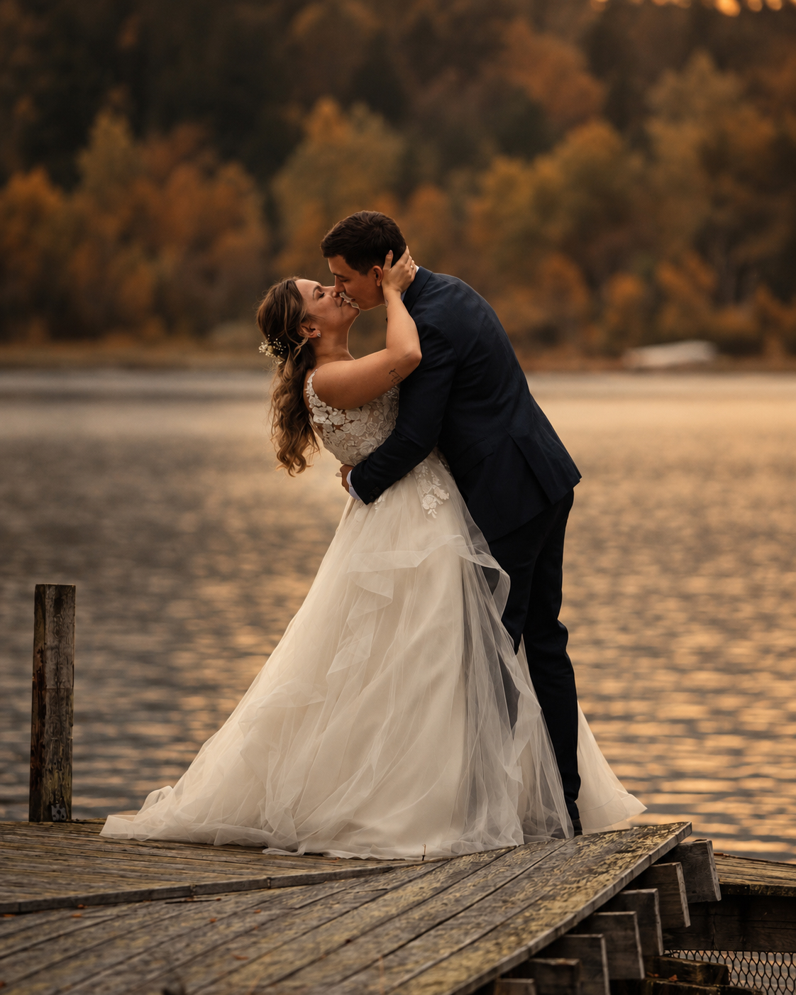 A bride and groom kissing on a wooden pier, the bride dipped backward in the groom's arms at sunset