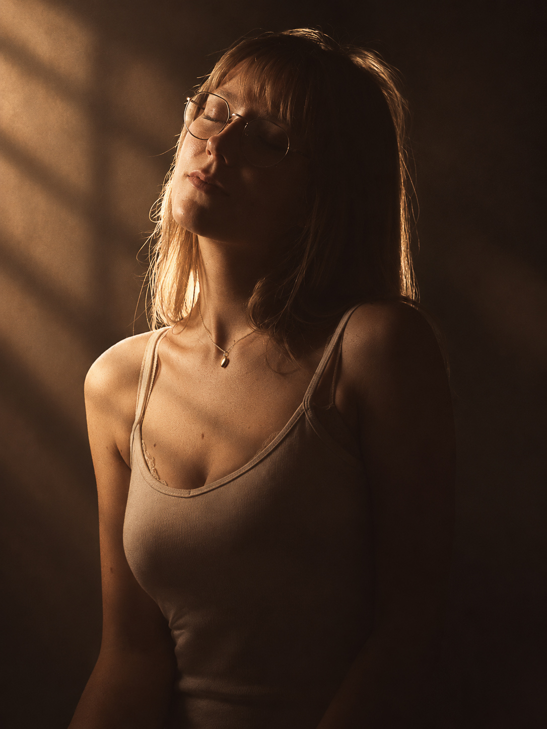 A young woman with long blonde hair and glasses, eyes closed in serene contemplation, lit by warm directional light with diagonal shadow patterns