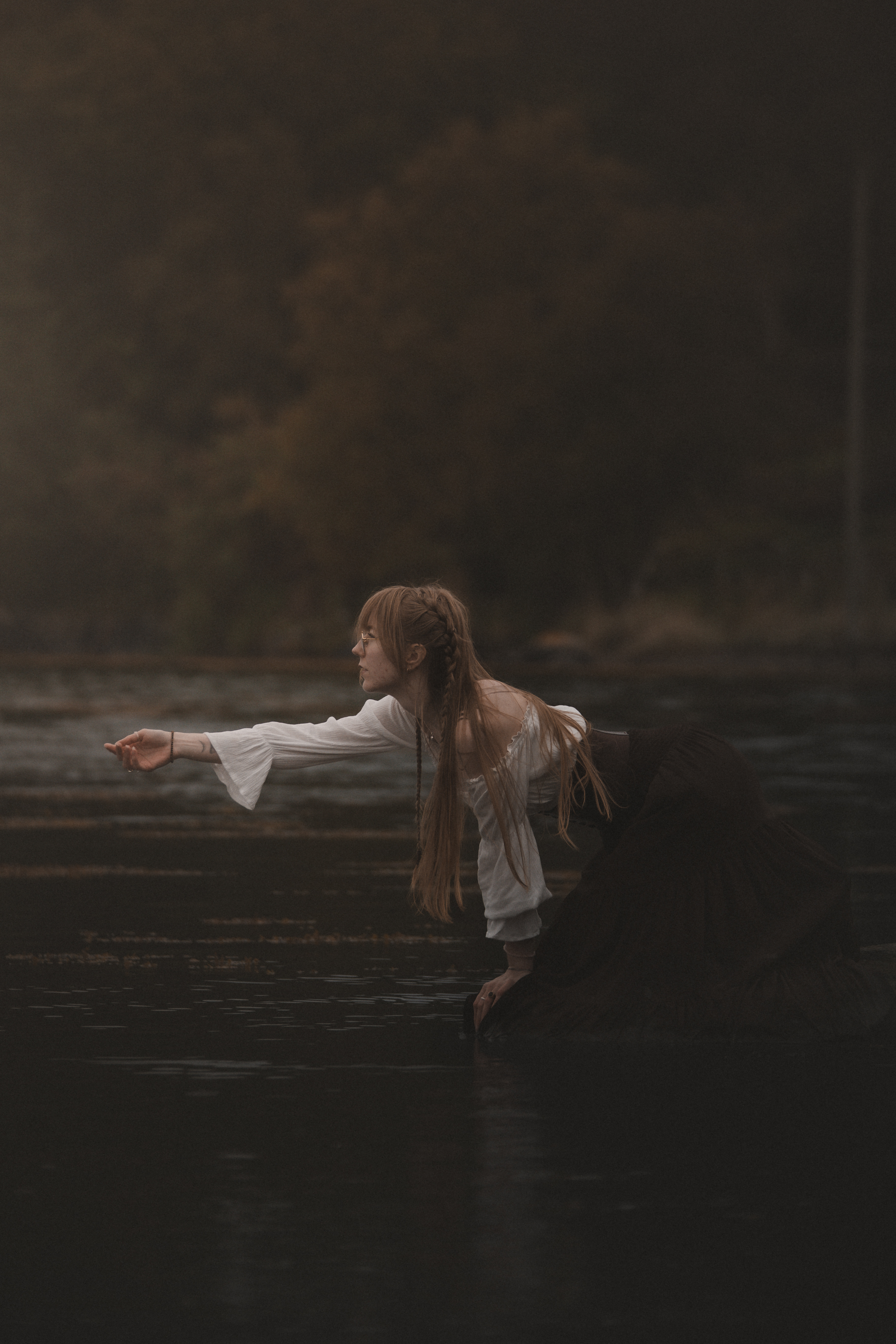 A young person with long braided reddish-brown hair kneeling in shallow dark water, reaching forward in profile
