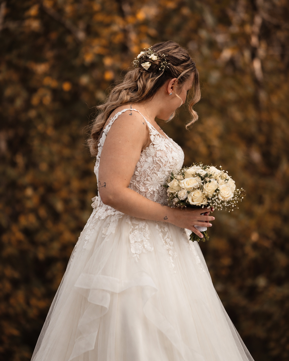 A bride in profile holding a bouquet of white roses, framed by soft autumn foliage